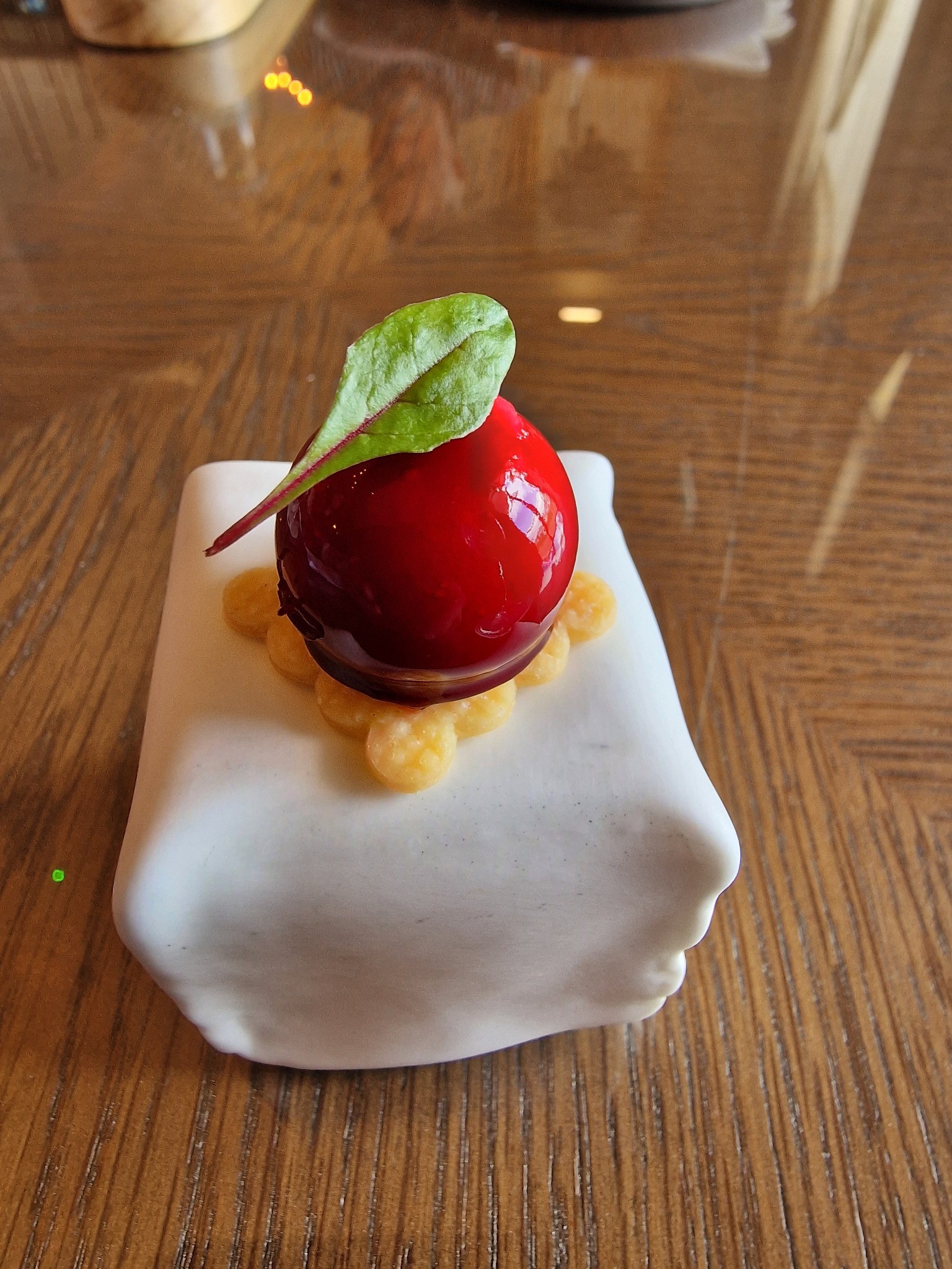 Square white dessert with a glossy red sphere and a green leaf on a wooden table