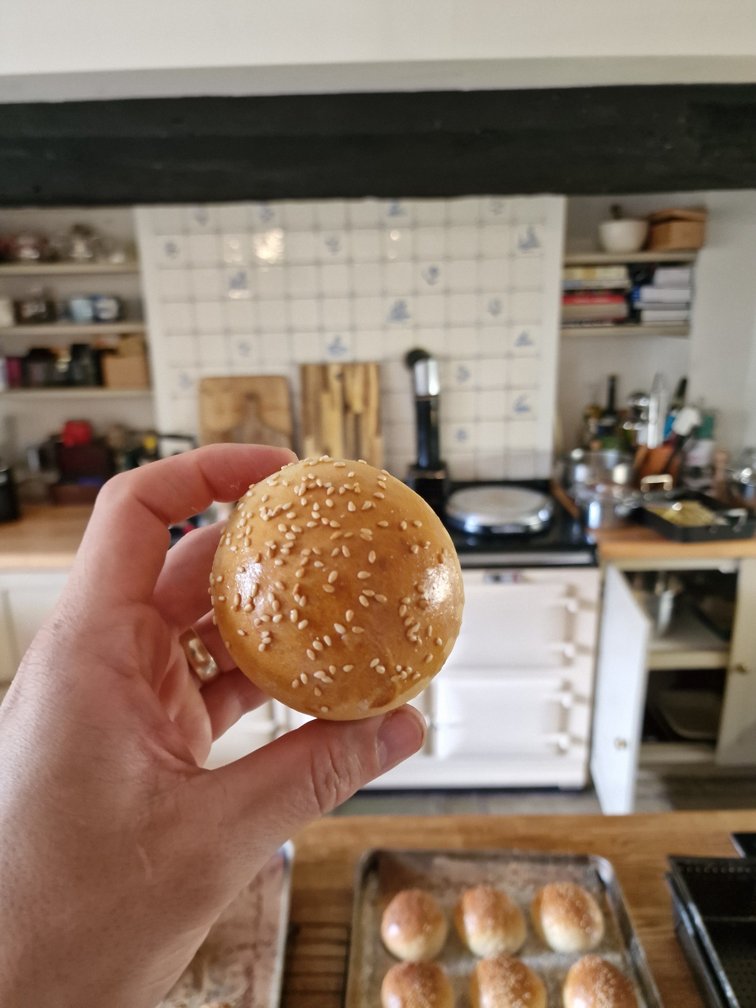 A hand holding a sesame seed bun with a tray of buns behind